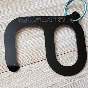 May include: Black plastic door opener with a loop for attaching to a keychain. The opener has a small loop on one end and a larger loop on the other end. The text "MePlusThee/ETSY" is engraved on the opener.