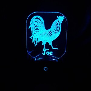 May include: A blue light illuminates a clear acrylic night light with a rooster design and the name "Joe" engraved on the bottom.