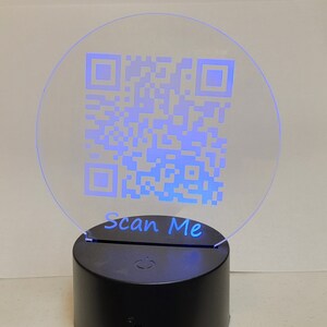 May include: A round, clear acrylic lamp with a blue light shining through it. The lamp has a QR code printed on it and the words "Scan Me" underneath. The lamp sits on a black base with a power button.