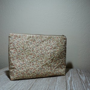 May include: A beige quilted zippered pouch with a floral pattern. The pouch is made of fabric and has a zipper closure.