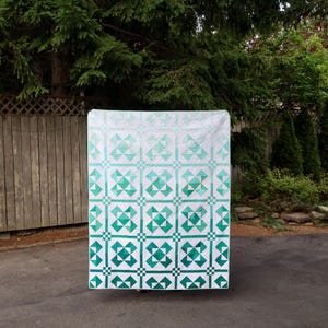 May include: A rectangular quilt featuring a repeating geometric pattern of squares and triangles in a gradient of green shades, transitioning from a light mint green at the top to a deep forest green at the bottom, set against a white background.