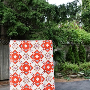 May include: A rectangular quilt featuring a repeating geometric pattern of large orange squares, light blue triangles, and smaller star-shaped blocks with brown and white floral prints on a white background, showcasing intricate machine-stitched quilting.