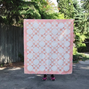 May include: A square quilt featuring a repeating geometric pattern of white circular shapes set against a soft peach-coloured background. The quilt has a wide peach border and displays intricate stitching throughout the entire fabric surface.