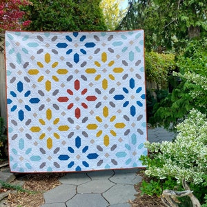 May include: A large quilt with a geometric pattern in shades of blue, yellow, red, and white. The quilt is hanging on a fence in a garden setting.