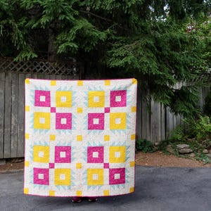 May include: A square quilt featuring a repeating geometric pattern of bright yellow and magenta squares arranged in a grid. The design includes light blue triangular accents and small yellow squares on a white background with a magenta border.
