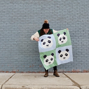 May include: A square quilt with four panda faces, each in a green or light blue square. The pandas have black ears, eyes, and patches, with a white face and a small black nose. The quilt is held up against a grey brick wall.