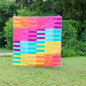 May include: A vibrant, geometric quilt in shades of pink, orange, yellow, teal, and purple. The quilt is displayed outdoors, held up against a backdrop of green foliage. The quilt's dimensions are not visible, but it is a striking piece.