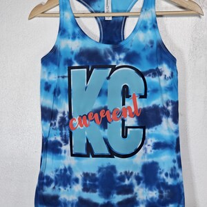 May include: A blue tie-dye tank top with a racerback design. The front features the letters "KC" in light blue with a black outline, and the word "current" in red script. The tank top is hanging on a wooden hanger.