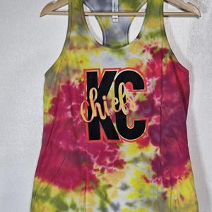 Könnte beinhalten: Ein Tanktop mit Batikmuster in Gelb, Rosa und Grün, das an einem Holzbügel hängt. Auf der Vorderseite des Tanktops steht der Text "KC Chiefs" in schwarzer und goldener Schrift. Das Tanktop hat ein Racerback-Design.