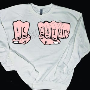 May include: White sweatshirt featuring two pink fist graphics. The left fist has the letters "KC" and the right fist has the letters "CHIEFS" in black. The sweatshirt is a Gildan brand.
