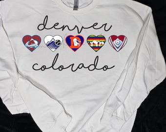 May include: White long-sleeved sweatshirt with the text "denver colorado" in black cursive lettering. Five heart-shaped logos are arranged horizontally above the text, each representing a different Denver sports team.
