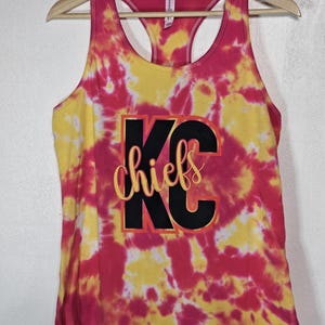Könnte beinhalten: Ein Tanktop im Batik-Design in Rosa und Gelb mit dem Text "KC Chiefs" in Schwarz und Gold. Das Racerback-Top wird auf einem Holzbügel präsentiert. Die Maße des Kleidungsstücks sind in Zentimetern.