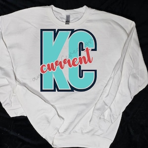 May include: White long-sleeved sweatshirt with a teal blue and navy blue "KC" graphic with the word "current" written in red on the front.