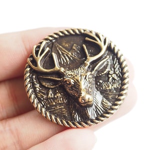 May include: A gold-toned metal pin featuring a detailed deer head design. The deer has large antlers and a textured, realistic face. The pin is surrounded by a rope-like border.