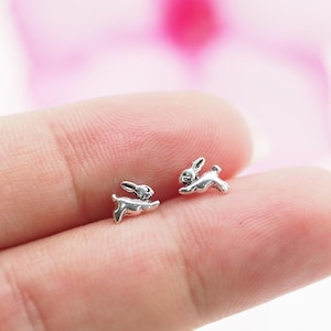 Op de afbeelding: Een paar kleine, zilverkleurige konijnenoorbellen. De oorbellen zijn in de vorm van rennende konijnen, met gedetailleerde kenmerken. De oorbellen worden op een vinger getoond, tegen een zachte, wazige roze en witte achtergrond.