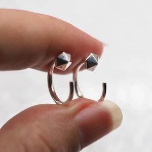 May include: A pair of silver hoop earrings. Each earring features a geometric, faceted design at the top of the hoop. The earrings are held between fingers against a soft, blurred background.