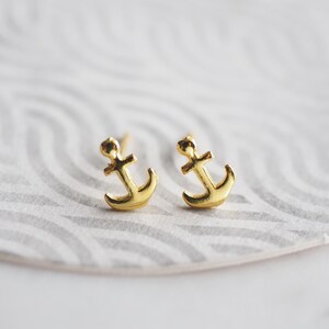 May include: A pair of gold-colored anchor stud earrings. The earrings are small, with a classic anchor design. The earrings are displayed on a white and gray patterned surface.