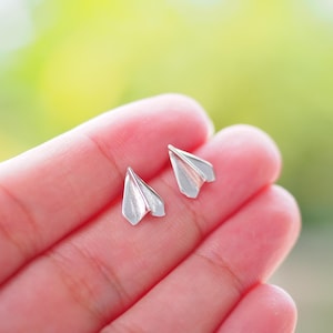 Può includere: Un paio di orecchini a forma di aeroplano di carta argentati. Gli orecchini sono piccoli e delicati, e sono perfetti per aggiungere un tocco di fantasia a qualsiasi outfit.