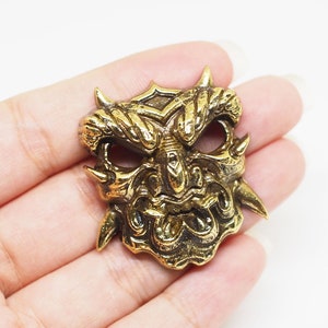 Könnte beinhalten: Eine detaillierte, goldfarbene Maske mit einem wilden Ausdruck, scharfen Hörnern und komplizierten Gesichtsdetails. Die Maske wird in einer Hand vor weißem Hintergrund gehalten und zeigt ihre Handwerkskunst und ihr Design.