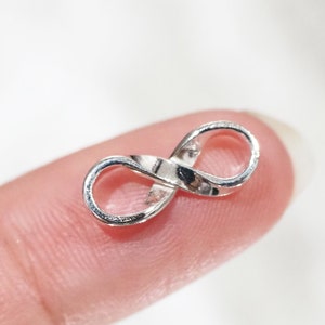 Pode incluir: Um pequeno pingente prateado com o símbolo do infinito. O pingente é segurado entre dois dedos, mostrando seu design delicado. O símbolo do infinito é um laço contínuo, representando a eternidade e as possibilidades infinitas.