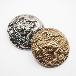 May include: Two round, decorative buttons with a raised dragon design. One button is silver and the other is gold.