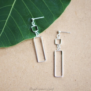 May include: Silver dangle earrings with a square and rectangle design. The earrings are on a brown surface with a green leaf.