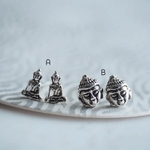 May include: Four silver earrings featuring a Buddha design. Two earrings feature a seated Buddha, and two earrings feature a Buddha head.