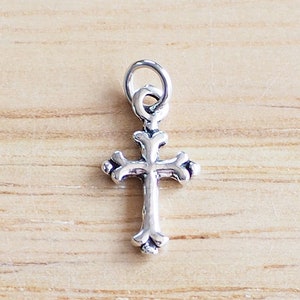 Può includere: Un piccolo ciondolo a forma di croce color argento con dettagli decorati. La croce presenta un anello nella parte superiore per appenderla ed è posta su uno sfondo marrone chiaro con venature del legno. Il ciondolo misura circa 2,5 cm di lunghezza.