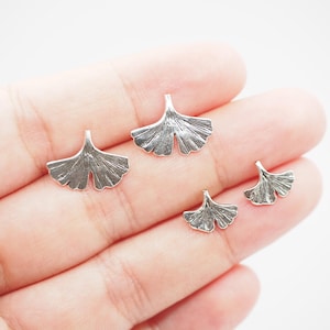 May include: A collection of silver-colored ginkgo leaf charms in various sizes. The charms have detailed veining and a textured finish, perfect for jewelry making or other craft projects. The leaves are displayed against a neutral background.
