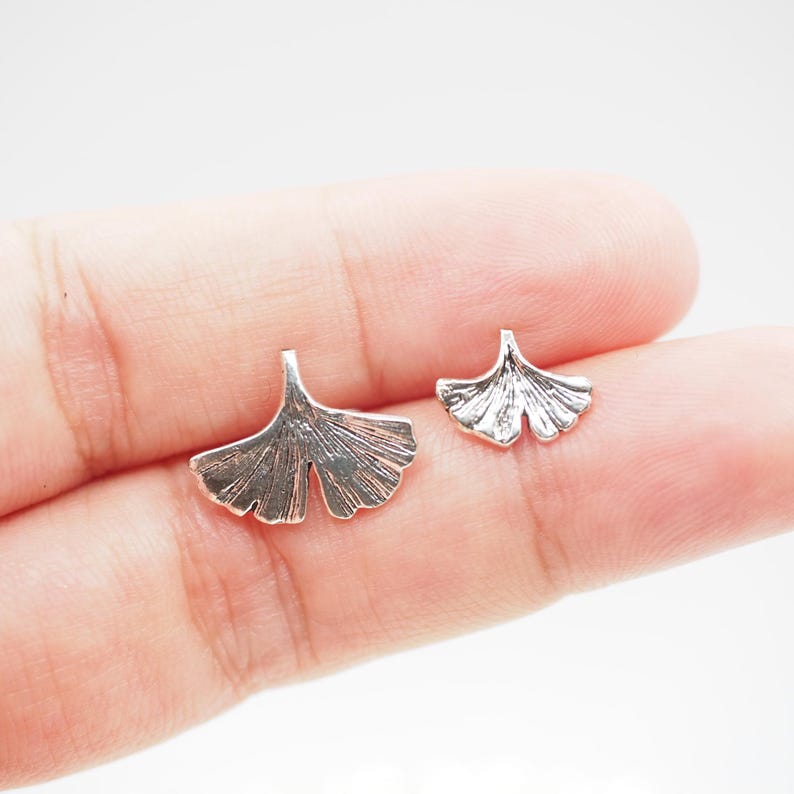 May include: Two silver-colored ginkgo leaf-shaped earrings. The earrings are detailed with veining and have a textured appearance. The leaves are displayed on a light background.
