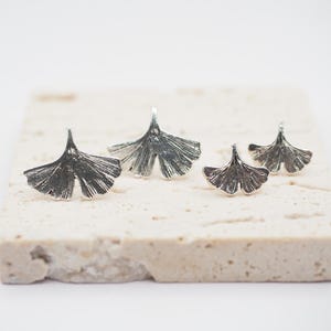 May include: Four silver-colored Ginkgo leaf-shaped jewelry pieces. The pieces are arranged on a light-colored, textured surface. The jewelry has detailed veining, and the leaves are slightly different sizes, suggesting earrings and a ring.