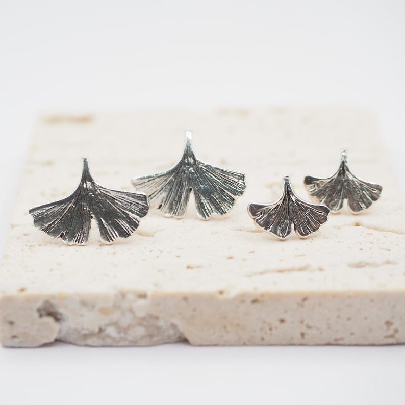 May include: Four silver-colored Ginkgo leaf-shaped jewelry pieces. The pieces are arranged on a light-colored, textured surface. The jewelry has detailed veining, and the leaves are slightly different sizes, suggesting earrings and a ring.
