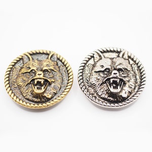 May include: Two circular metal buttons featuring a raised relief of a snarling wolf head in the centre. Each button is framed by a decorative rope-style border. One button has a warm brass finish, while the other has a bright silver finish.