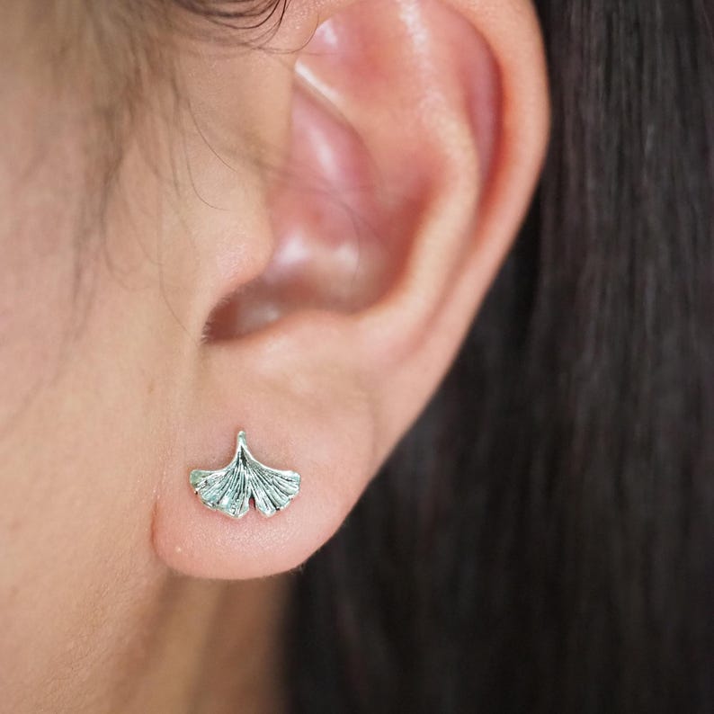 May include: A close-up of a silver-colored stud earring in the shape of a ginkgo leaf. The earring is detailed with fine lines and is worn on an ear. The background is blurred, focusing attention on the jewelry.