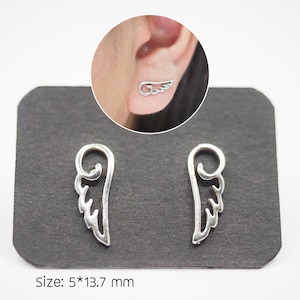 Puede incluir: Un par de pendientes plateados con forma de ala. Los pendientes se muestran sobre una superficie gris oscuro. El tama&ntilde;o de los pendientes es de 5 mm por 13,7 mm. Un primer plano muestra un pendiente en uso.