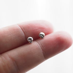May include: A pair of small, silver-colored stud earrings, each shaped like a tiny, textured skull. The earrings have a metallic, reflective finish and are presented as individual pieces, showcasing their detailed, irregular surface and compact size.