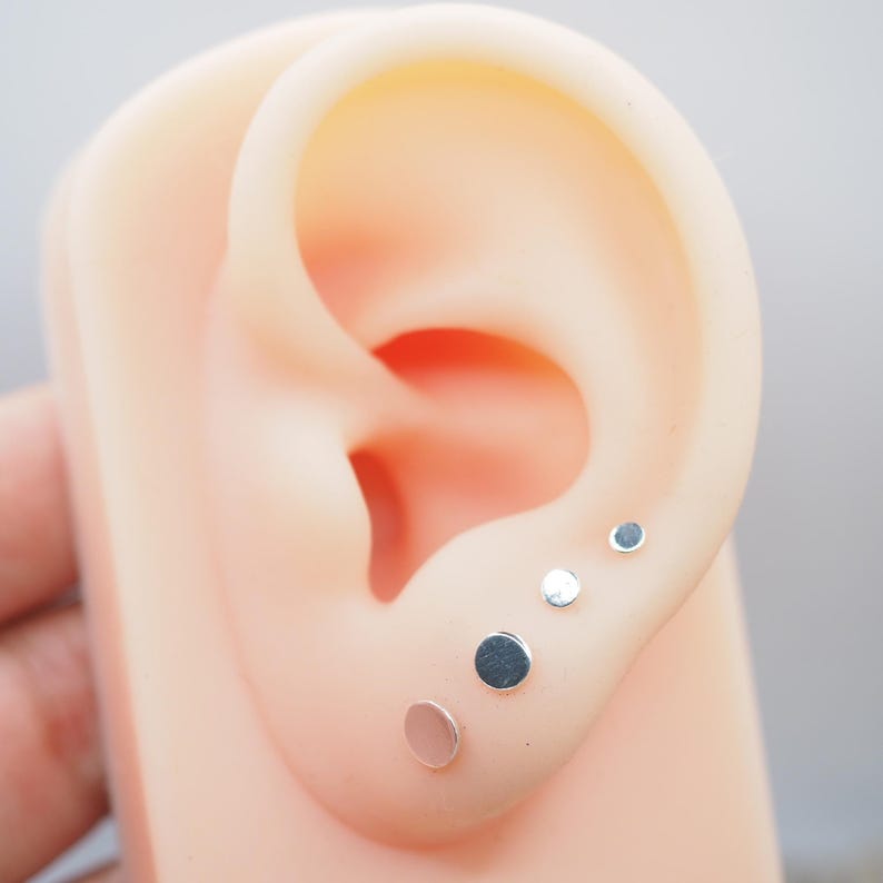 May include: Five silver stud earrings of different sizes are shown in a close-up image of a model ear.