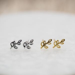 Op de afbeelding: Twee paar kleine, delicate oorbellen. Het ene paar is zilver en het andere is goud. Beide paren hebben een eenvoudig, bladvormig ontwerp.