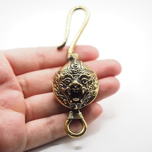 May include: A gold-toned brass hook featuring a central, spherical ornament embossed with a detailed, snarling mythical creature face. The object includes a curved top hook for suspension and a small circular ring at the base for attachment.