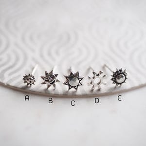 Op de afbeelding: Vijf zilveren neusringen met verschillende zonontwerpen. De ontwerpen omvatten een zon met een gezicht, een zon met stralen, een zon met een cirkel in het midden en twee verschillende zonontwerpen met stralen.