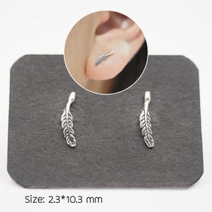 Puede incluir: Un par de pendientes de pluma plateados sobre una superficie gris oscura. Los pendientes miden aproximadamente 2,3 mm por 10,3 mm. Un primer plano muestra un pendiente en uso. El texto "Size: 2.3*10.3 mm" es visible.