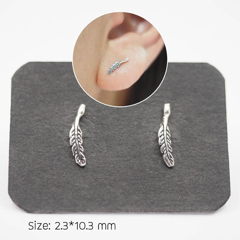 Puede incluir: Un par de pendientes de pluma plateados sobre una superficie gris oscura. Los pendientes miden aproximadamente 2,3 mm por 10,3 mm. Un primer plano muestra un pendiente en uso. El texto "Size: 2.3*10.3 mm" es visible.