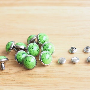 May include: A group of green and silver metal rivets with a marbled design. The rivets are arranged in a pile on a wooden surface. There are also four silver metal rivets with no design on the surface.