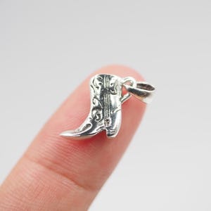May include: Silver charm in the shape of a boot with a floral design. The charm is on a finger.