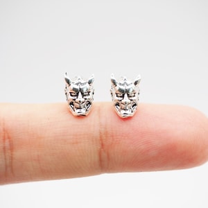 Può includere: Un paio di orecchini d'argento con un design dettagliato di maschera oni. Le maschere hanno un'espressione feroce con corna e denti aguzzi.