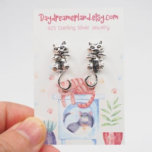 Op de afbeelding: Een paar zilveren kattenoorbellen op een witte kaart met een kattenillustratie. De kaart heeft de tekst "Daydreamerland.etsy.com 925 Sterling Silver Jewelry".