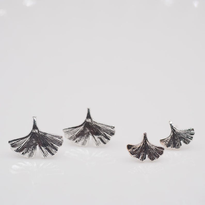 May include: Four silver-colored ginkgo leaf earrings. The earrings are arranged in a group, with two larger pairs and two smaller pairs. The leaves have detailed veining and a textured appearance, set against a white background.
