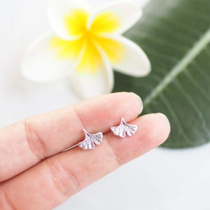 May include: A pair of silver-colored stud earrings in the shape of ginkgo leaves. The earrings are displayed on a hand, with a white and yellow flower and a green leaf in the background.