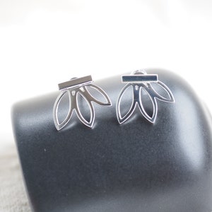 May include: A pair of silver-colored earrings with a floral design. Each earring features three petal-shaped cutouts below a rectangular bar. The earrings are displayed on a dark gray surface against a white background.