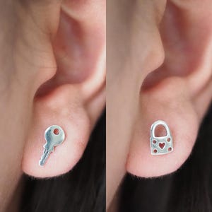 May include: Silver key and lock stud earrings. The key earring is on the left ear, and the lock earring is on the right ear. The lock earring has a heart-shaped cutout.